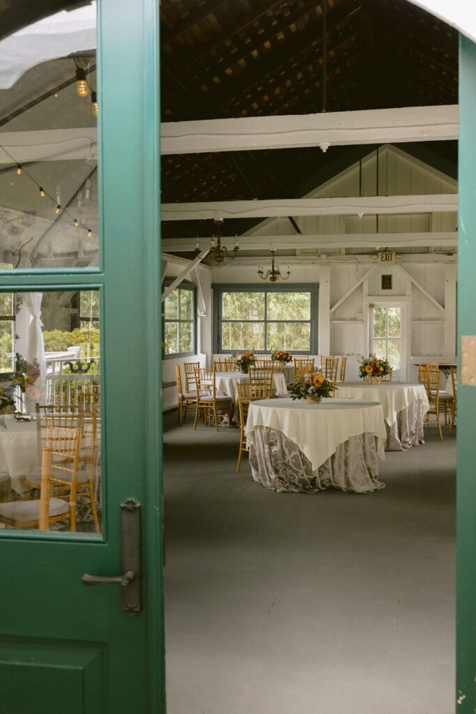A green door opens to a rustic event hall with round tables covered in white cloths, floral centerpieces, and wooden chairs. String lights and vintage chandeliers add warmth to the bright, airy space.