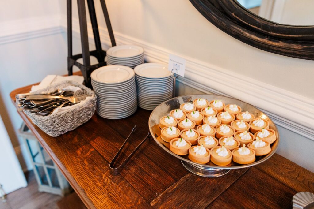 A wooden table with stacked white plates, a basket of utensils, and a tray of small tarts topped with whipped cream. Tongs rest on the tray, and a round mirror is visible on the wall.