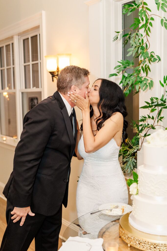 A bride in a white dress kisses her groom in a black suit beside a white wedding cake, while standing near a window with greenery decorations.