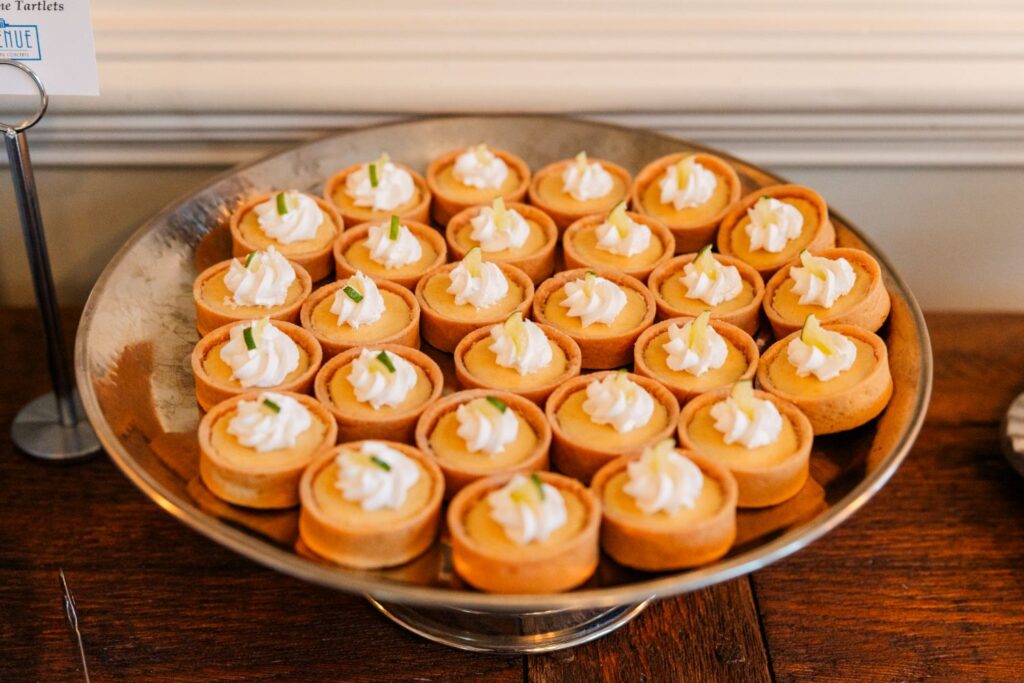 A tray of mini tartlets with golden crusts, filled with creamy filling, topped with a swirl of whipped cream and small green garnish, displayed on a wooden table.