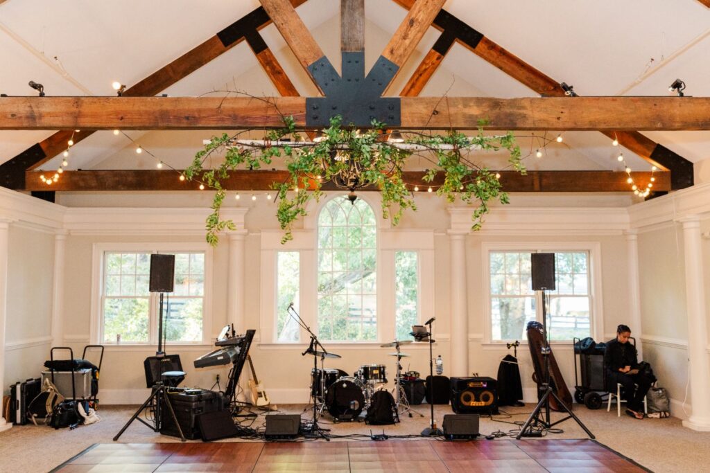 A wedding band setup with instruments on a stage under wooden beams and hanging greenery, with string lights and large windows in the background.