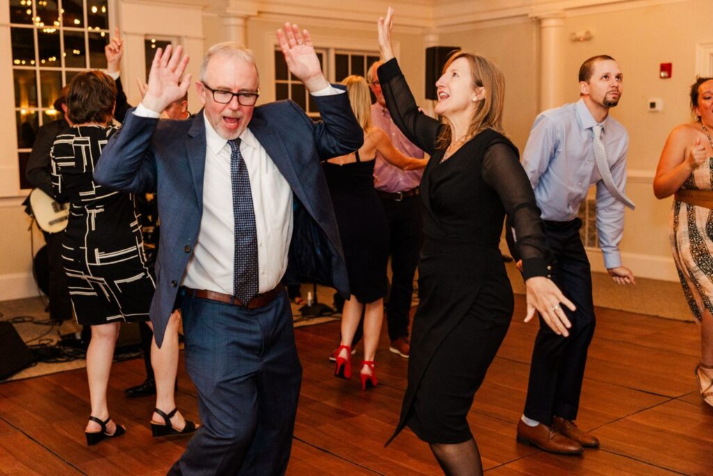 A group of people dance energetically on a wooden floor at a formal event, smiling and raising their hands in the air. The atmosphere is lively and joyful, with festive attire and warm lighting.