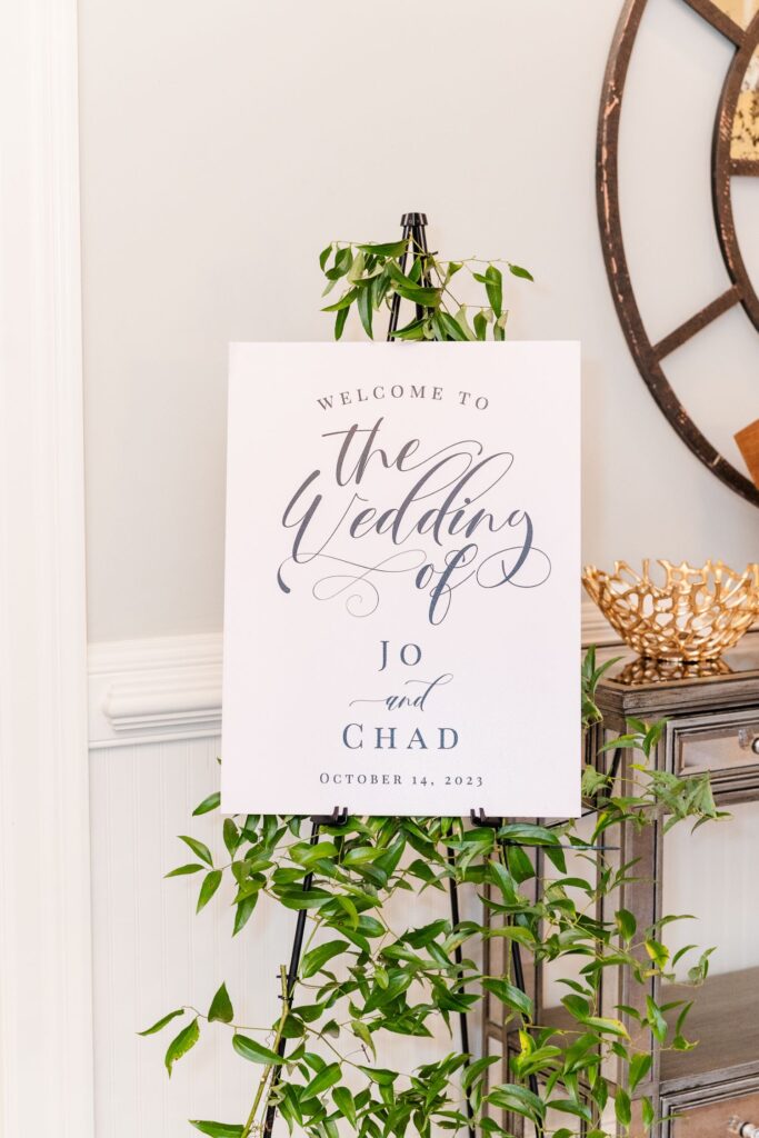 A white sign on an easel with greenery reads, Welcome to the Wedding of Jo and Chad, October 14, 2023, in elegant script, next to a table and partial clock.