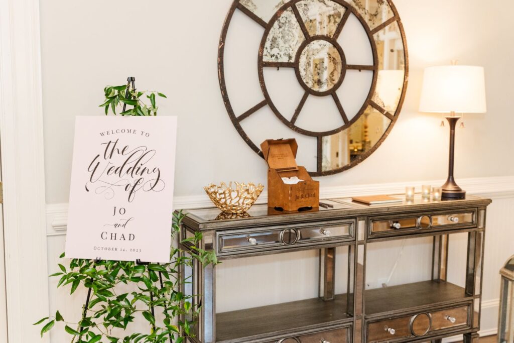 A wedding welcome sign for Jo and Chad is displayed next to a decorative table with a mirror, lamp, gold bowl, and wooden box in a warmly lit room.