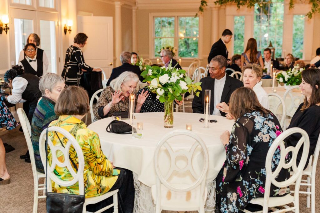 A group of people sits around a round table at a formal event. The table is decorated with flowers and candles. Some guests are chatting and smiling, while staff serve in the background.