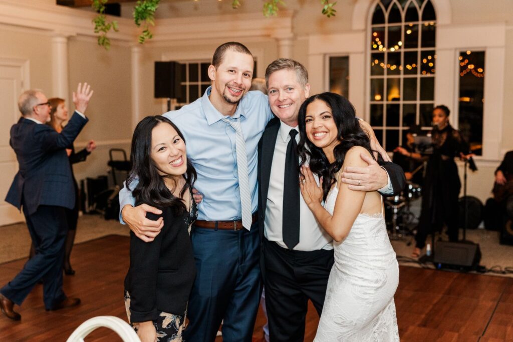 Four people pose and smile together at a wedding reception, with two men and two women, one in a white dress. Guests and musicians are in the background of the warmly lit room.