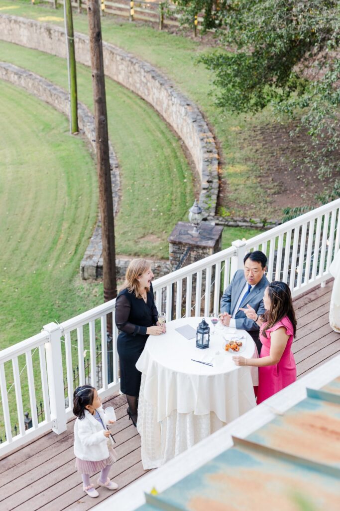 Four adults stand around a tall, round table on a patio, talking and eating. A young girl in a white jacket walks nearby. The patio overlooks a grassy area bordered by a stone wall and wooden fence.