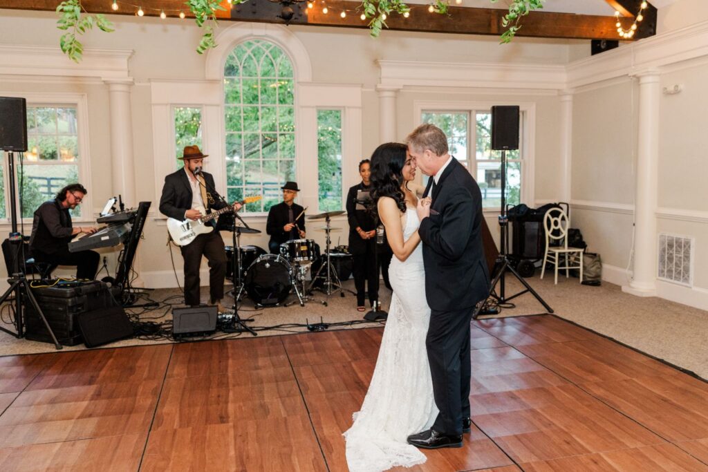 A bride and groom share their first dance on a wooden floor as a live band performs in the background of a bright, elegant room with large windows.