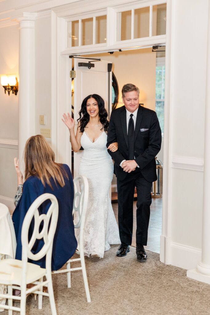 A woman in a white dress and a man in a suit walk through a doorway, smiling. The woman waves while guests seated at white chairs look toward them.