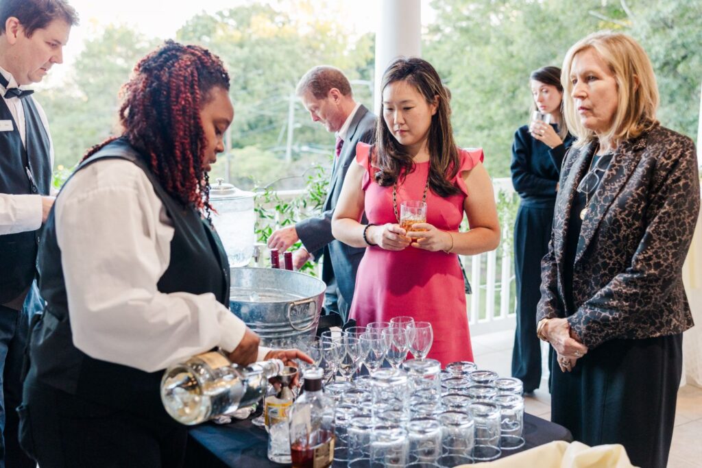 A bartender pours a drink at a table lined with glassware while three guests, including a woman in a pink dress, wait and watch during a daytime event on a porch.