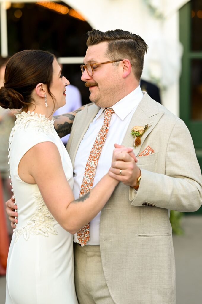 A couple dressed in wedding attire shares a joyful dance outdoors, smiling at each other. The bride wears a white dress with lace details, and the groom wears a light suit with a patterned tie.