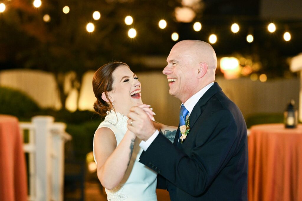 A woman in a white dress and a man in a suit are laughing and dancing together outdoors at night under string lights.