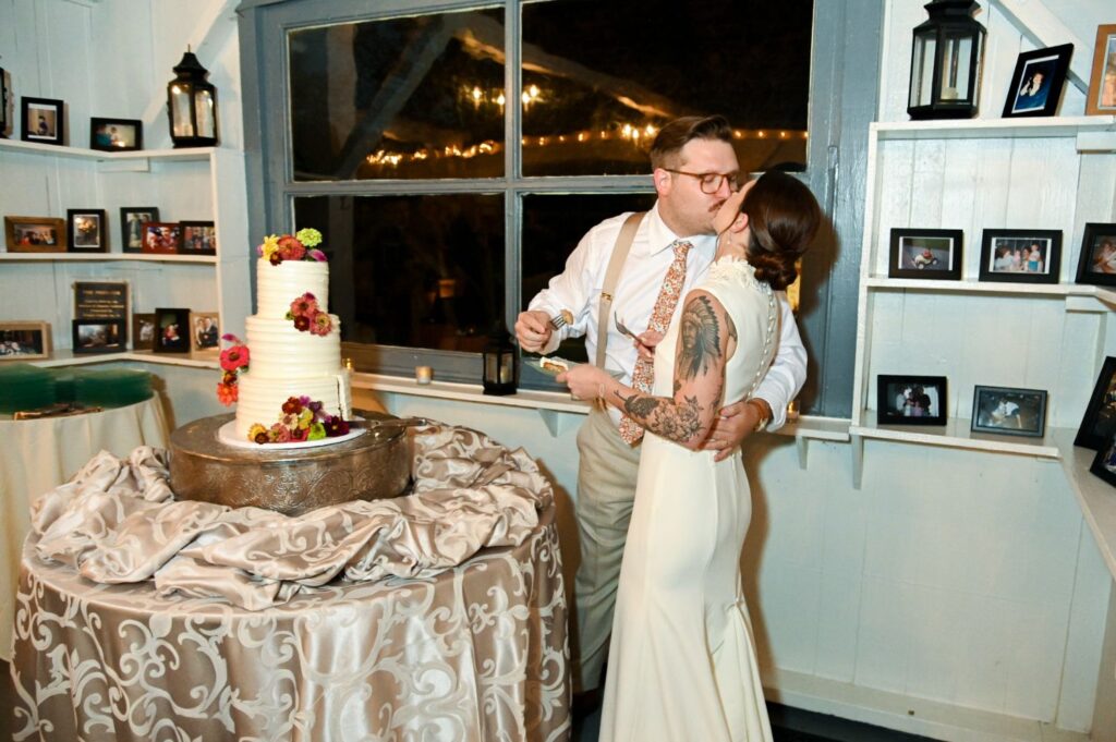 A bride and groom share a kiss next to their wedding cake in a cozy room decorated with photos and soft lighting.