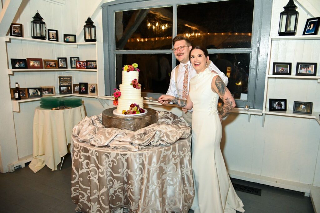 A couple in wedding attire smiles while cutting a white tiered cake with colorful flowers. The cake sits on a round table with a patterned cloth. Shelves with framed photos and lanterns are in the background.