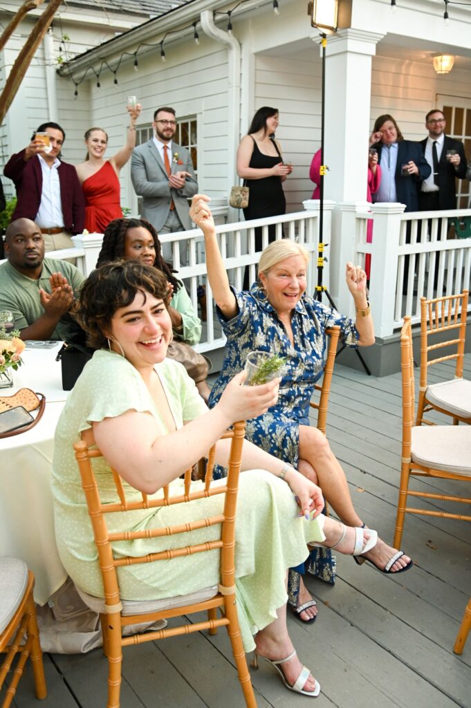 A group of people at an outdoor event raises their glasses in a celebratory toast. Some are seated at a table, others stand on a porch behind them, all smiling and engaged in the moment.