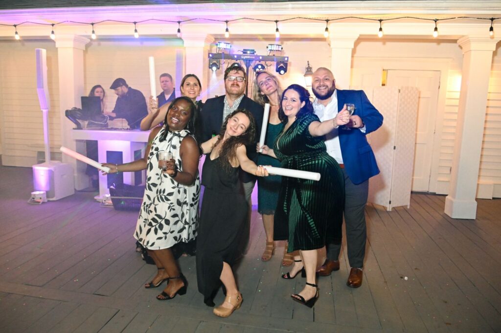 A group of people dressed up, smiling and posing for a photo at a party with string lights above and DJs in the background. Some hold light sticks and one person holds a drink.