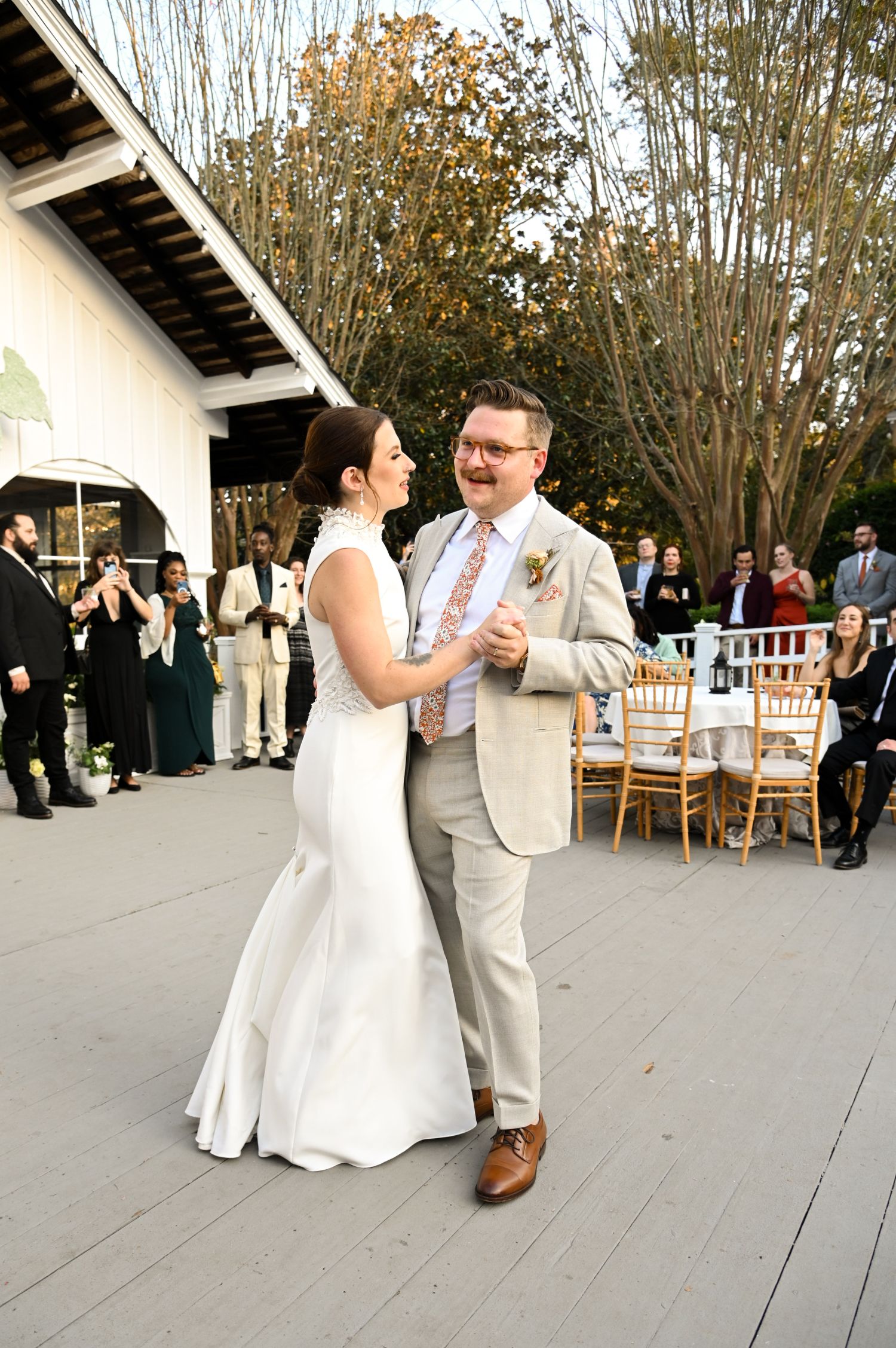 A bride and groom smile and dance together outdoors at their wedding reception, surrounded by guests watching and taking photos.