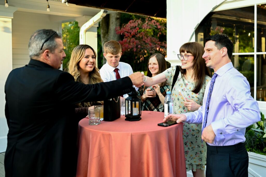Six people stand around a round table with a coral tablecloth, smiling and talking outdoors. Drinks and a lantern sit on the table, and greenery is visible in the background.
