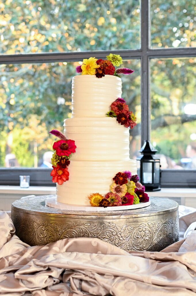 Three-tiered white cake decorated with colorful flowers sits on an ornate silver stand, with a window and greenery in the background.