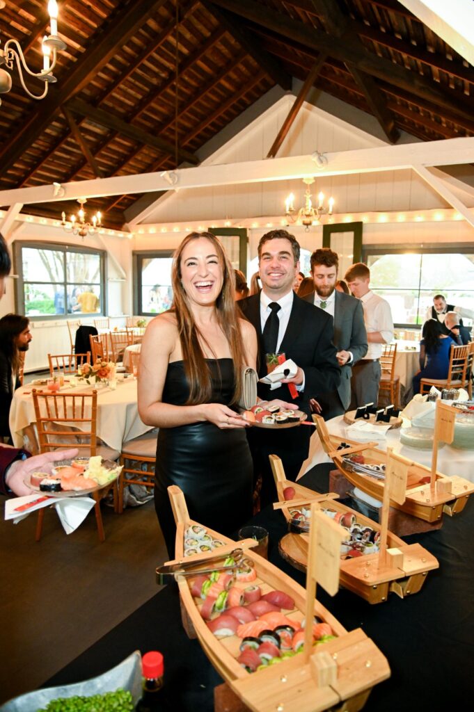 A woman in a black dress and a man in a suit smile while serving themselves sushi at a buffet table during an indoor event with wooden chairs and string lights.