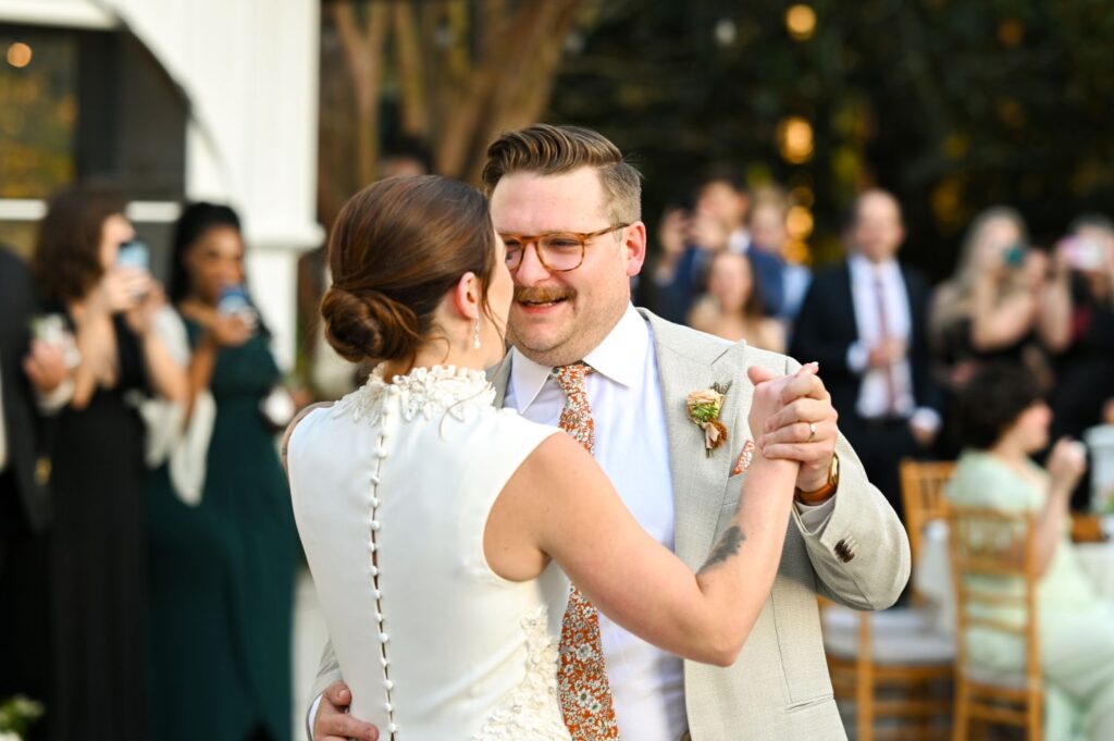 A bride and groom share their first dance outdoors, surrounded by guests who watch and take photos. The groom wears glasses and a suit, while the bride wears a white dress with buttons down the back.