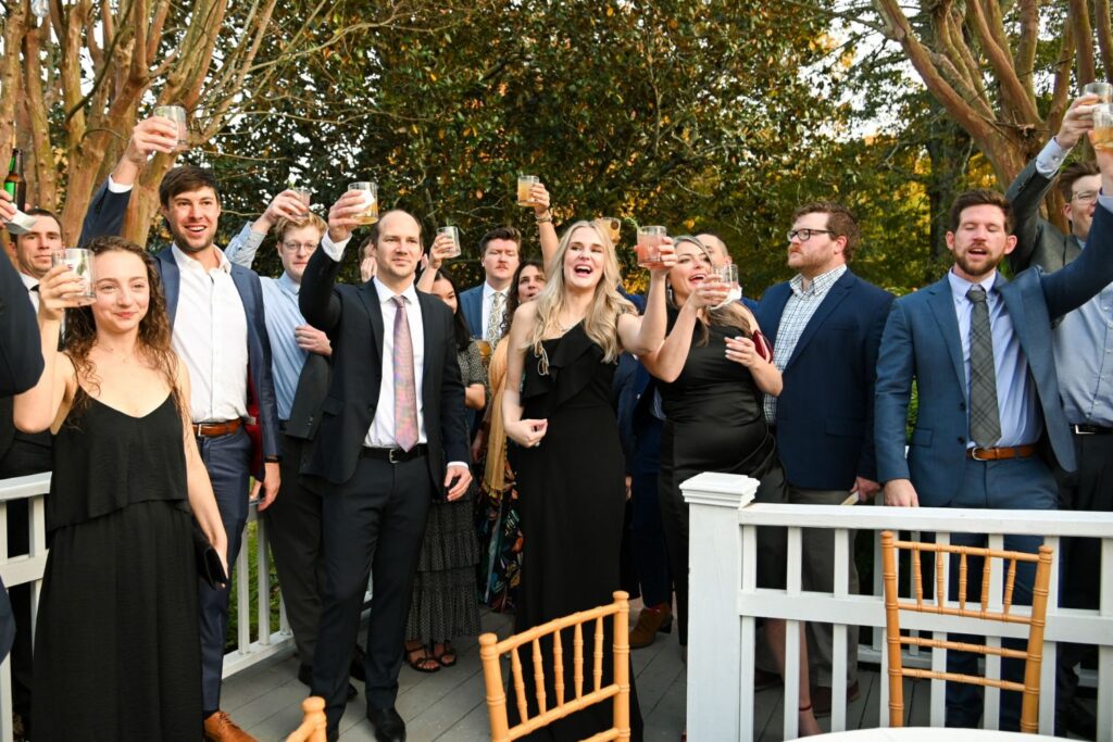 A group of people dressed in formal attire stand on a deck, raising glasses in a celebratory toast. Trees and greenery are in the background, and empty chairs are in the foreground.