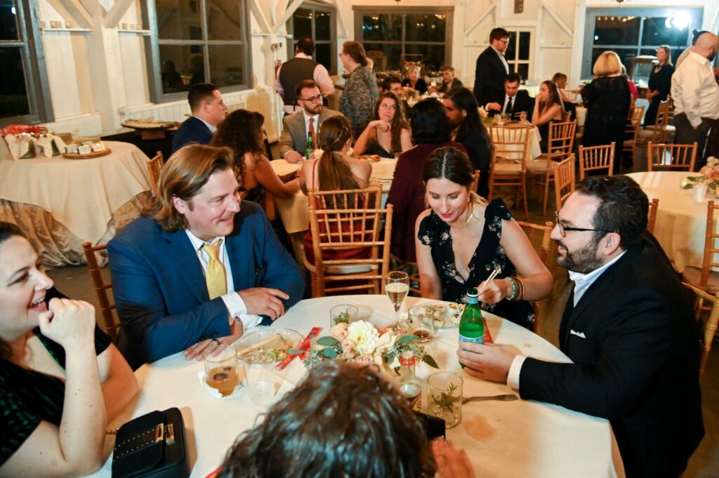 People in formal attire sit around round tables at a lively indoor event, eating, drinking, and conversing. The background shows more guests mingling and dining, with warm lighting creating a festive atmosphere.