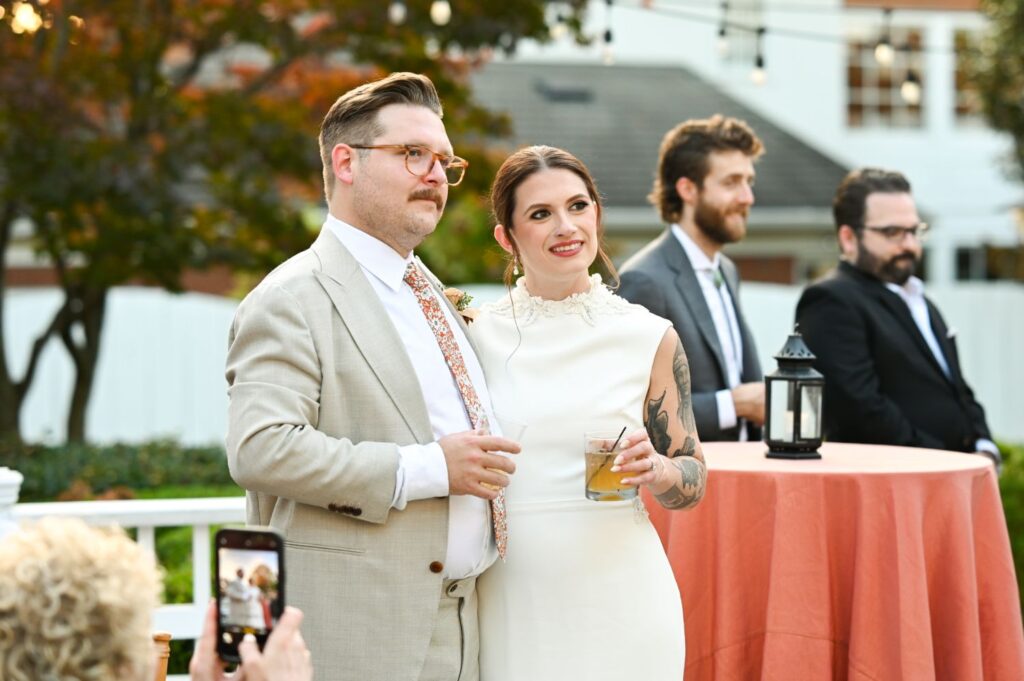A bride and groom stand together outdoors, smiling and holding drinks. The bride wears a white dress, the groom a beige suit. Two guests stand by a round table with a coral tablecloth in the background. String lights hang above.