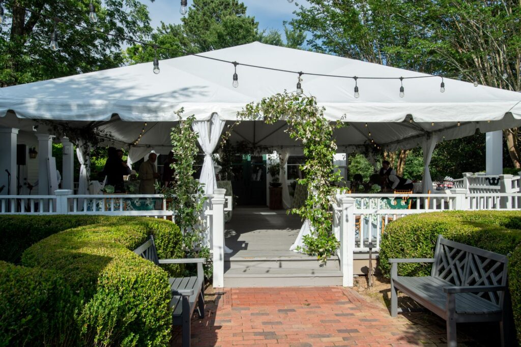A white event tent decorated with greenery stands on a wooden deck, with benches and trimmed bushes in front. String lights hang overhead, creating a charming ambiance, and guests can be seen inside the tent on a sunny day—an ideal setting for those searching for unique engagement party venues in Atlanta.