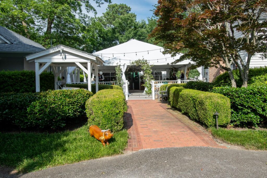 A brick walkway leads to a white tented patio surrounded by lush green bushes and trees, creating an inviting atmosphere perfect for engagement party venues Atlanta. There is a small orange cow sculpture on the left side of the path, while string lights hang above the patio, adding a charming glow to the scene.