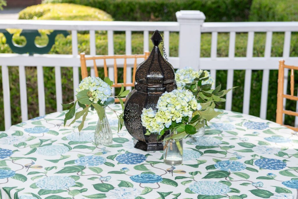 A round table with a floral tablecloth holds three small vases of blue hydrangeas and a decorative black lantern. Set outdoors with a white fence and greenery in the background, this charming setup beautifully captures the inviting ambiance found at top engagement party venues Atlanta has to offer.