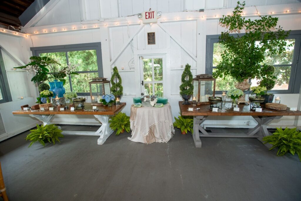 A decorated room with two wooden tables holding plants, flowers, and food displays offers an inviting setting reminiscent of top engagement party venues Atlanta has to offer. A small round table with a white cloth and green plates is centered between them. Ferns sit on the floor, and windows reveal greenery outside.