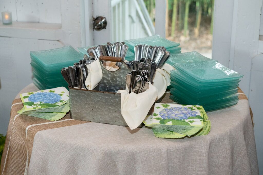 A round table holds stacks of green glass plates, floral napkins with blue hydrangeas, and a metal container filled with silverware wrapped in white napkins—a charming setup often found at the most inviting engagement party venues Atlanta has to offer.