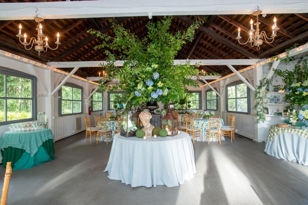 A decorated event space with wooden beams, chandeliers, round tables covered in white cloth, and a central table featuring a large floral arrangement and statues. Greenery and natural light fill the room, making it an elegant choice among engagement party venues Atlanta has to offer.