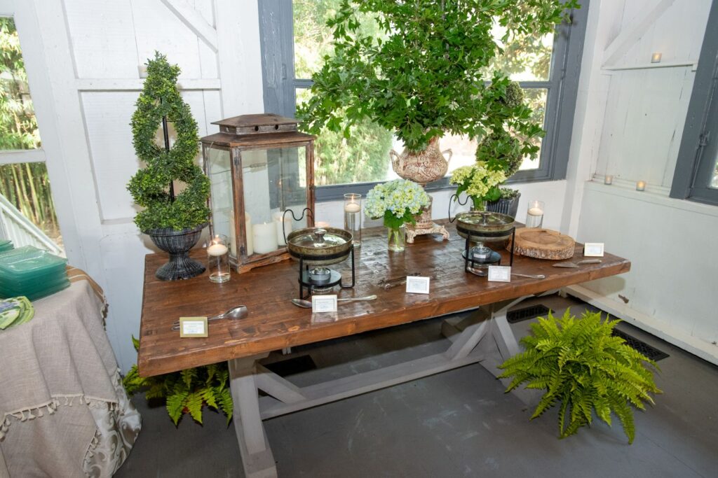 A rustic wooden table holds chafing dishes, candles, cards, plants, and flowers. The setting is bright with natural light, greenery, and decorative accents—creating an elegant buffet display reminiscent of the sophisticated ambiance found at top engagement party venues Atlanta has to offer.