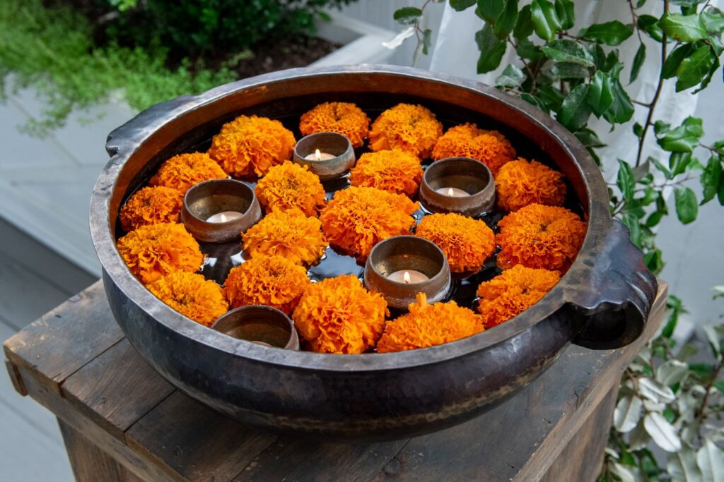 A large metal bowl filled with water, floating orange marigold flowers, and small clay oil lamps, placed on a wooden surface with green plants in the background—this elegant arrangement is a perfect decorative inspiration for engagement party venues in Atlanta.