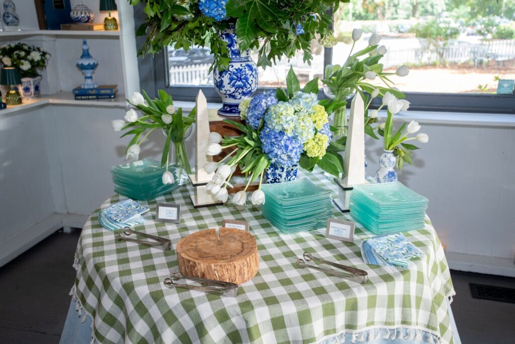 Round table with green plaid cloth, green glass plates, blue floral napkins, tongs, and white name cards. Floral arrangements with white tulips and blue hydrangeas decorate the table, creating an elegant setting ideal for those seeking engagement party venues Atlanta has to offer. Shelves with vases are in the background.