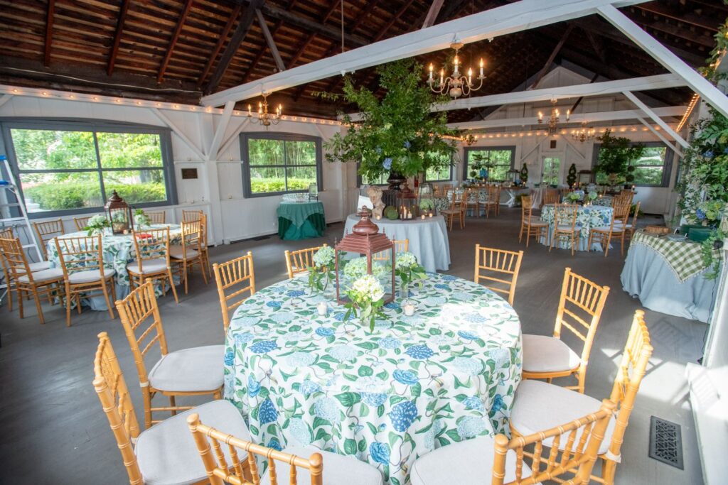 A bright, rustic event hall with round tables covered in floral tablecloths, wooden chairs, chandeliers, potted plants, and large windows letting in natural light—one of the most charming engagement party venues Atlanta has to offer.