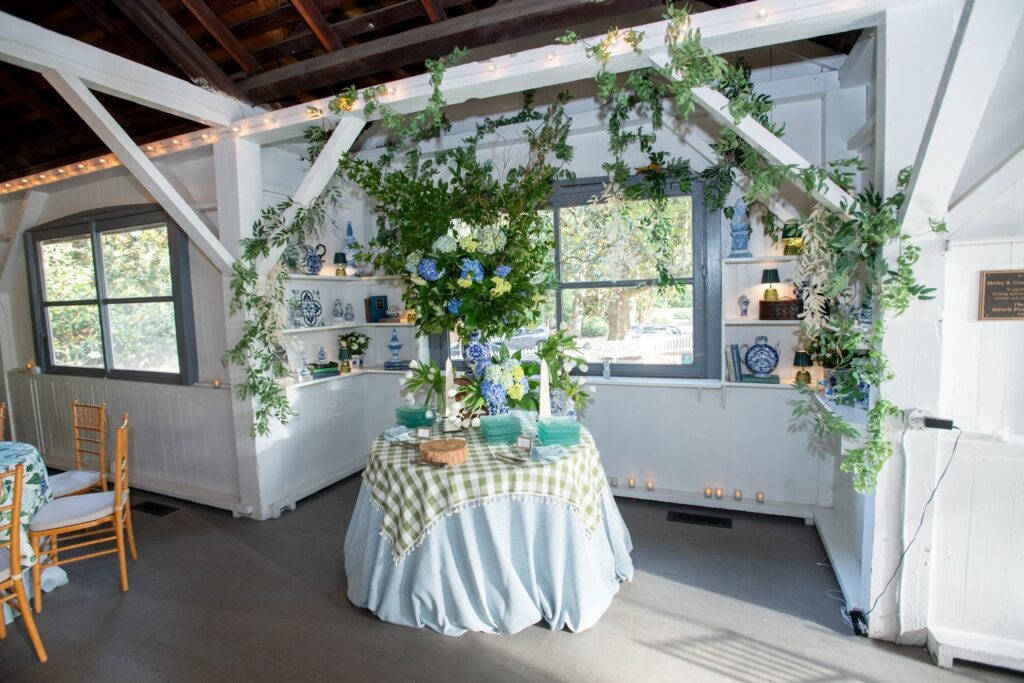 A round table with a green checkered cloth holds blue and white vases, greenery, and glassware. Reminiscent of some of the most charming engagement party venues Atlanta has to offer, the white room is decorated with leafy garlands, shelves of ceramics, and string lights, with windows letting in daylight.