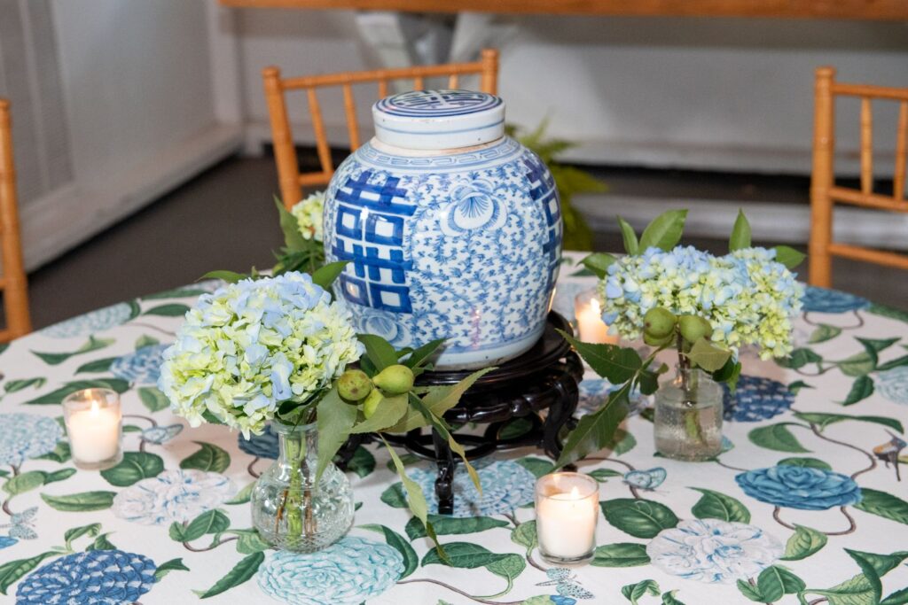 A blue and white ceramic jar sits on a stand, surrounded by small bouquets of hydrangeas and four lit candles on a floral tablecloth with blue and green flower patterns—an elegant display that would grace the finest engagement party venues Atlanta has to offer.