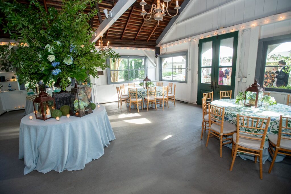 Bright, airy event space with round tables covered in blue cloths, wooden chairs, floral centerpieces, candles, and large windows letting in natural light. Perfect for those seeking engagement party venues Atlanta offers, this location features a large floral arrangement on a draped round table to elevate your celebration.