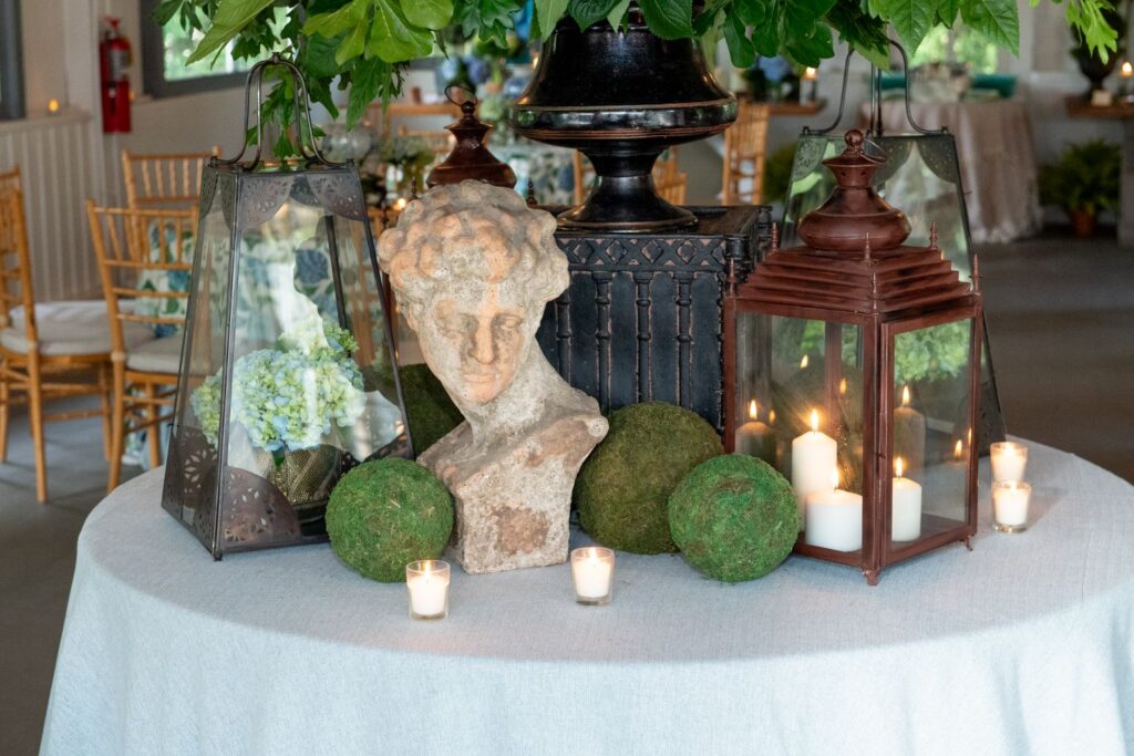 A round table with a white cloth holds a classical bust, two lanterns with candles, green moss balls, and small votive candles, with leafy greenery above—creating an elegant atmosphere reminiscent of top engagement party venues Atlanta has to offer. Wooden chairs and more tables are visible in the background.