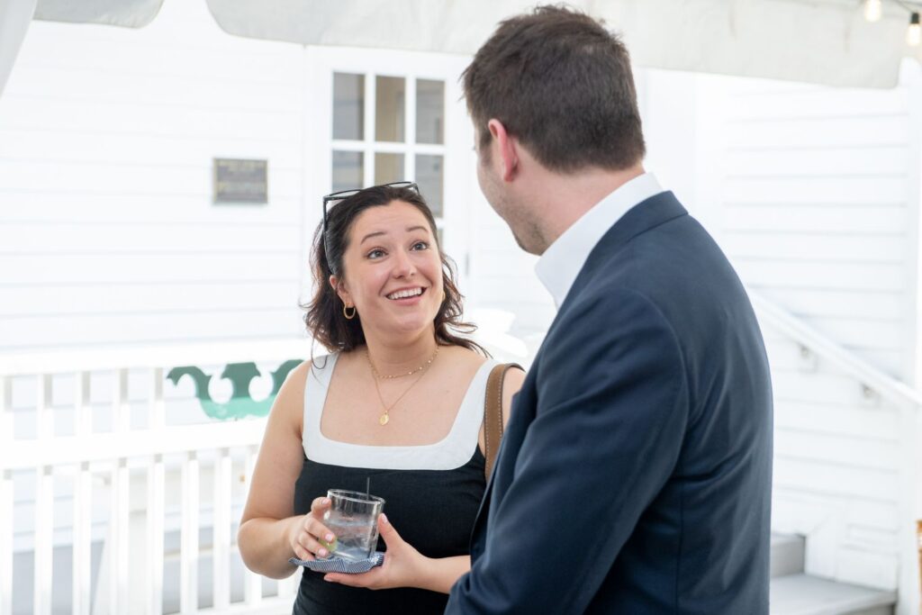A woman holding a drink smiles and talks with a man in a suit at an outdoor event with a white building in the background, showcasing one of the elegant engagement party venues Atlanta has to offer.