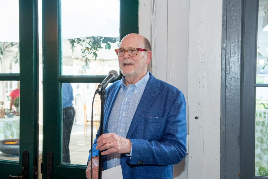 An older man in a blue blazer and glasses speaks into a microphone indoors, standing near a window with green trim, holding a piece of paper in his left hand—a scene that could easily unfold at one of the many elegant engagement party venues Atlanta has to offer.