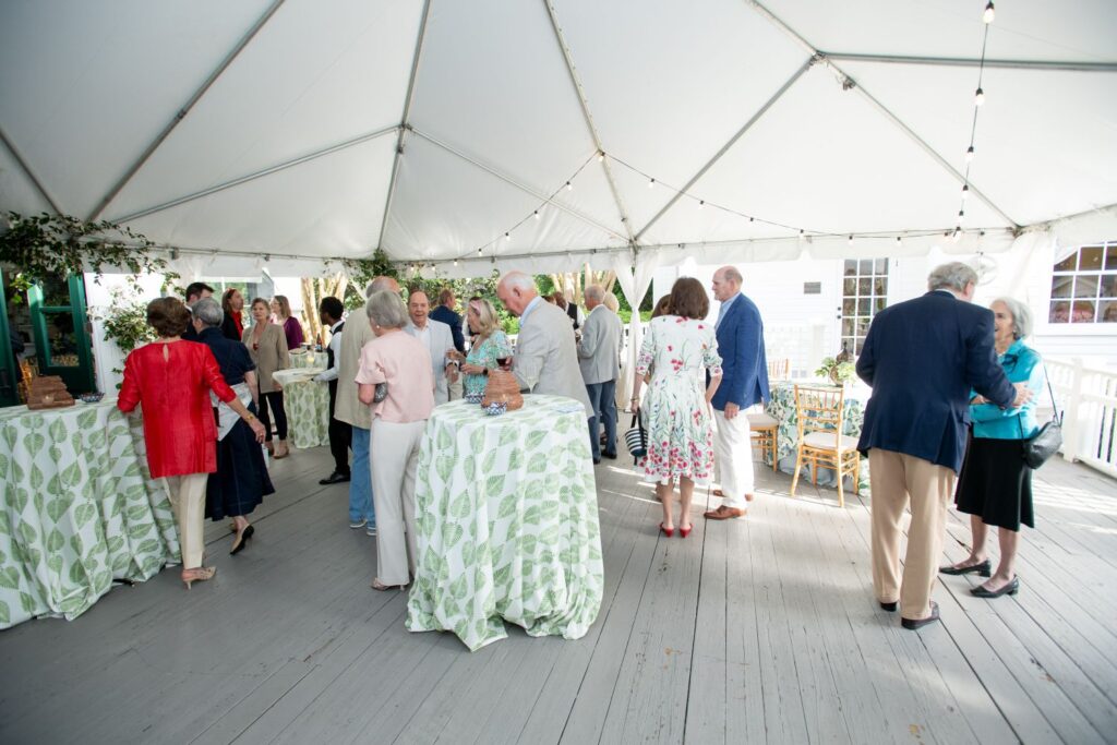 A group of people dressed in semi-formal attire socialize under a white event tent, standing around tables covered with green leaf-patterned tablecloths. String lights are visible above, creating an inviting atmosphere perfect for those seeking unique engagement party venues Atlanta has to offer.