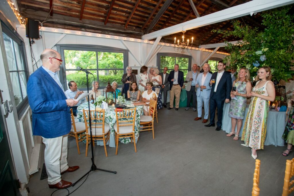 A man in a blue jacket speaks at a microphone to a group of people dressed in semi-formal attire gathered inside one of the most charming engagement party venues Atlanta has to offer, featuring elegant décor, wooden beams, and a sparkling chandelier.