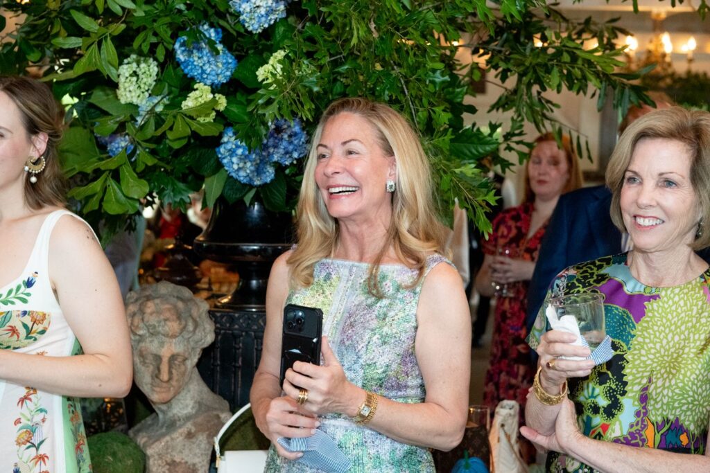 Three women smiling and holding drinks at a lively indoor gathering, surrounded by lush green foliage and blue hydrangeas—an atmosphere reminiscent of the best engagement party venues Atlanta has to offer. One woman holds a phone and cloth mask while people mingle in the background, creating a warm and inviting celebration.