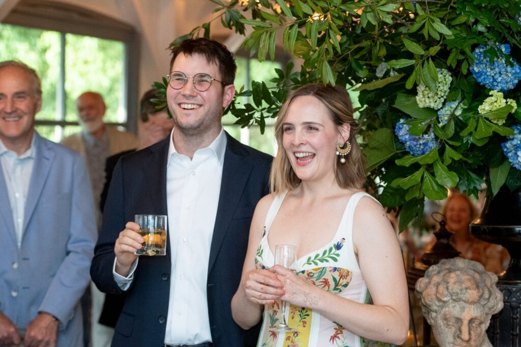 A smiling man in a suit and a woman in a floral dress hold drinks at a lively indoor gathering, surrounded by guests and lush green floral decor—perfect inspiration for those searching for elegant engagement party venues Atlanta has to offer.