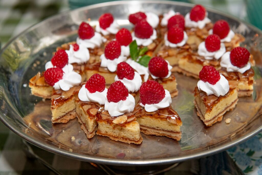 Triangular dessert bars topped with whipped cream, raspberries, and a glaze are arranged on a round silver platter. Perfect for celebrations hosted at engagement party venues Atlanta is known for, the treats stand out against a green checked tablecloth in the background.