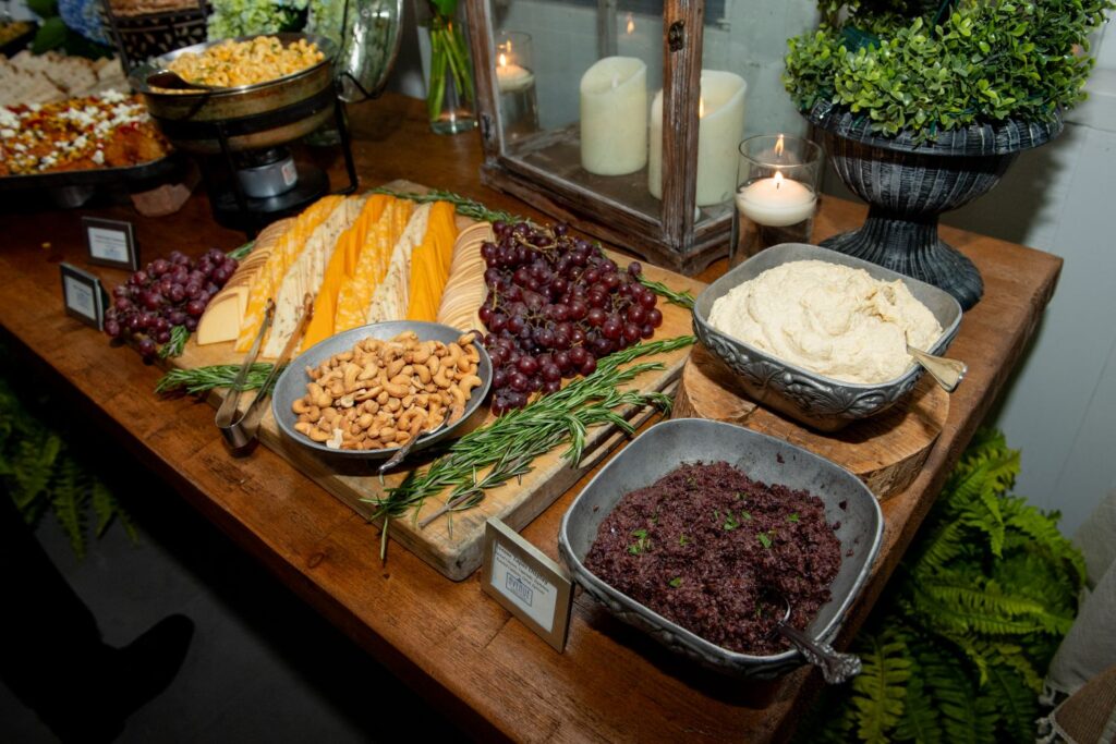 A wooden table with assorted cheeses, red grapes, rosemary, mixed nuts, hummus, tapenade, and candles is beautifully arranged for a buffet or appetizer spread—an inviting setup perfect for those seeking inspiration for engagement party venues in Atlanta. Green plants decorate the edges of the table, adding a fresh and elegant touch to the display.